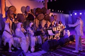 A group of musicians dressed in traditional attire, playing instruments and singing on a stage. The musicians in the front row are seated and hold sheet music, while those standing behind are playing large hand drums. The stage is decorated with white curtains and jars, with lighting equipment visible.