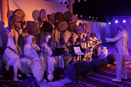 A group of musicians dressed in traditional attire, playing instruments and singing on a stage. The musicians in the front row are seated and hold sheet music, while those standing behind are playing large hand drums. The stage is decorated with white curtains and jars, with lighting equipment visible.
