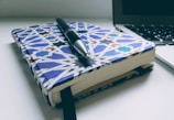 A notebook with an intricate geometric pattern cover lies on a desk alongside a pen and a partially visible laptop. The cover design features blue, white, and other colors in a symmetrical layout.