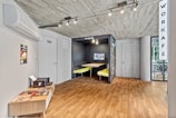 The image depicts an interior space with a modern and minimalist design. A small area labeled 'TV Room' features a built-in booth with yellow seats and a wall-mounted television. The floor is a light wood laminate, and the ceiling is unfinished concrete. On the left, there is an air conditioning unit above a small table that holds magazines. A door with a sign is next to a wall poster showing a dish. In the background, a glass wall separates the area from another room labeled 'Workafe'.