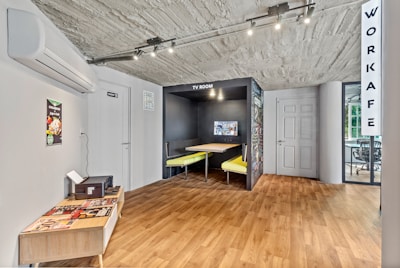 The image depicts an interior space with a modern and minimalist design. A small area labeled 'TV Room' features a built-in booth with yellow seats and a wall-mounted television. The floor is a light wood laminate, and the ceiling is unfinished concrete. On the left, there is an air conditioning unit above a small table that holds magazines. A door with a sign is next to a wall poster showing a dish. In the background, a glass wall separates the area from another room labeled 'Workafe'.