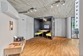 The image depicts an interior space with a modern and minimalist design. A small area labeled 'TV Room' features a built-in booth with yellow seats and a wall-mounted television. The floor is a light wood laminate, and the ceiling is unfinished concrete. On the left, there is an air conditioning unit above a small table that holds magazines. A door with a sign is next to a wall poster showing a dish. In the background, a glass wall separates the area from another room labeled 'Workafe'.