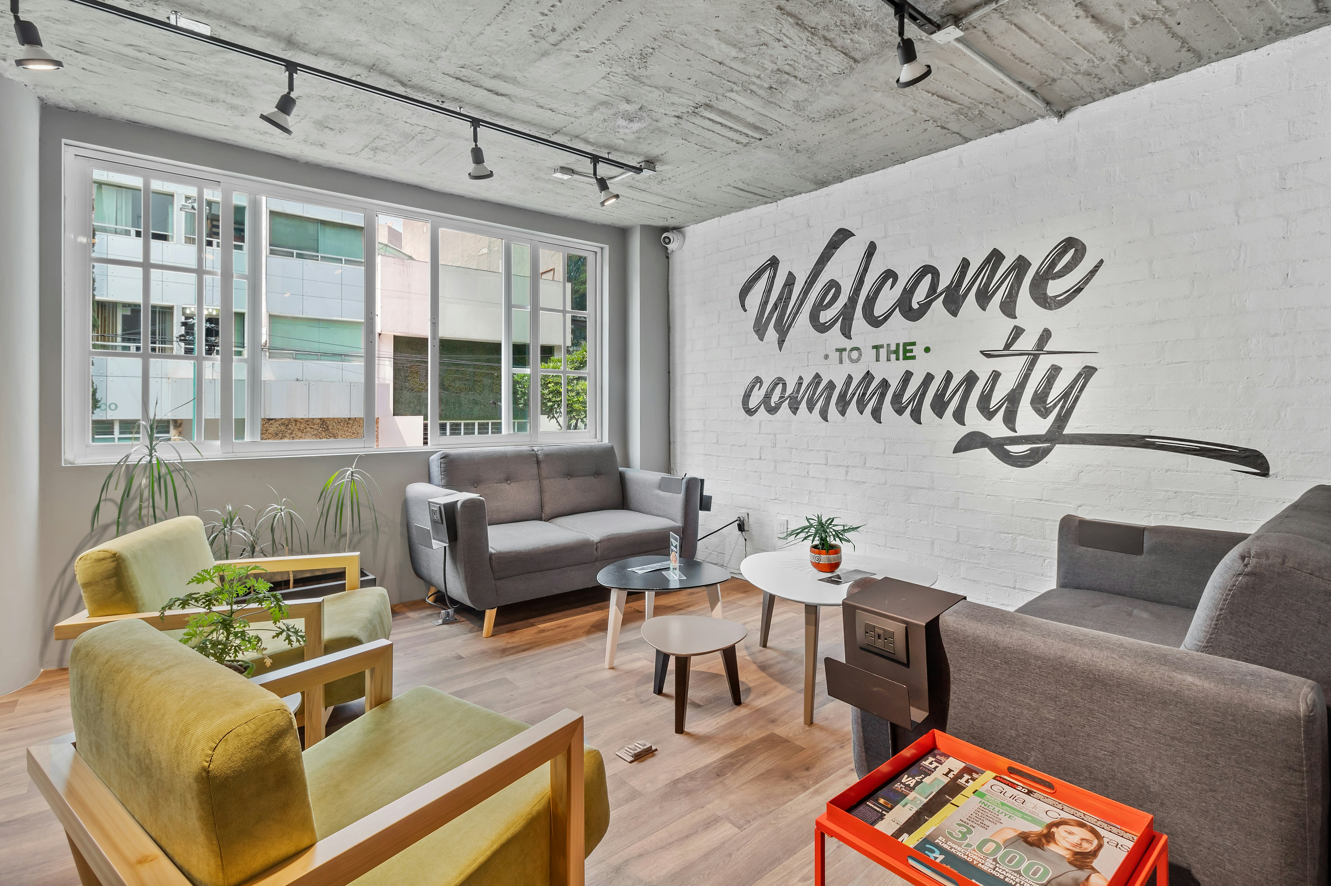 A room with hardwood floors and grey and geen chairs and loveseats.  The wall has painted words that says Welcome to the community.