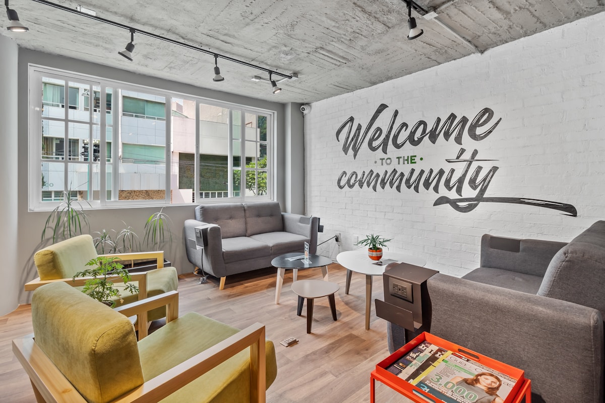 A room with hardwood floors and grey and geen chairs and loveseats.  The wall has painted words that says Welcome to the community.