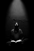 A person sitting cross-legged, reading a book in a dimly lit room. Light shines down from above, illuminating the person's face and the open book. The background is a rough, textured wall.