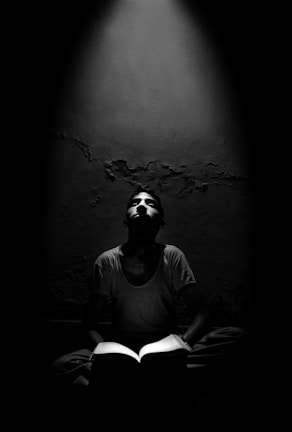 A person sitting cross-legged, reading a book in a dimly lit room. Light shines down from above, illuminating the person's face and the open book. The background is a rough, textured wall.