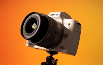 A DSLR camera mounted on a tripod, set against a vibrant orange background. The lens is prominently visible, and the camera body is angled, showcasing its design.