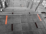 Stairway with metal railings on both sides, featuring gray tiled steps and flooring. Orange directional arrows are marked on the tiles, suggesting movement or guidance. The geometric patterns create a structured and orderly aesthetic.