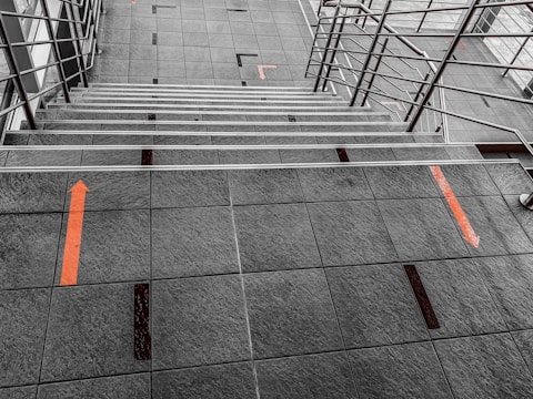Stairway with metal railings on both sides, featuring gray tiled steps and flooring. Orange directional arrows are marked on the tiles, suggesting movement or guidance. The geometric patterns create a structured and orderly aesthetic.