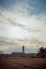 A tall brick building with a cross on top, possibly a church or school, is set against a cloudy sky. The structure is surrounded by a large grassy area with some trees nearby.