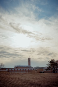 A tall brick building with a cross on top, possibly a church or school, is set against a cloudy sky. The structure is surrounded by a large grassy area with some trees nearby.