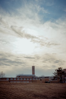 A tall brick building with a cross on top, possibly a church or school, is set against a cloudy sky. The structure is surrounded by a large grassy area with some trees nearby.