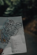 A hand holding a folded map with detailed pathways and labeled locations. The map features various symbols and a legend, which includes a list of destinations. The background is blurry, suggesting a natural setting with trees.