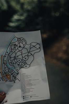A hand holding a folded map with detailed pathways and labeled locations. The map features various symbols and a legend, which includes a list of destinations. The background is blurry, suggesting a natural setting with trees.