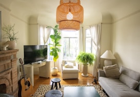 A cozy living room with a combination of modern and rustic decor. There is a large wicker light fixture hanging from the ceiling, casting warm light. The space features a brick fireplace on the left, with decorative items on the mantel. A flat-screen TV and a guitar are positioned near the fireplace. In front of a large window with sheer curtains, several potted plants, including a large fern and a fiddle leaf fig, add greenery to the room. The seating area includes a cushioned armchair with a pillow and a gray sectional sofa, accompanied by a white lamp. A small animal print rug and a trio of dark, round nesting tables complete the setup on the wooden floor.