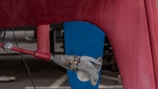 A close-up view of a boat propeller attached to the bottom of a red hull. The propeller is metallic and appears slightly weathered. The background includes a blue vertical structure and a concrete surface.