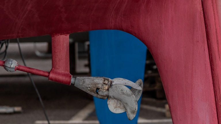 A close-up view of a boat propeller attached to the bottom of a red hull. The propeller is metallic and appears slightly weathered. The background includes a blue vertical structure and a concrete surface.