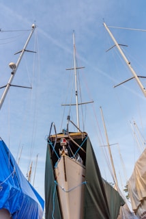 Several sailboats are docked and covered with tarps and sheets. The masts tower high into a clear blue sky, and various ropes are used to secure the coverings.