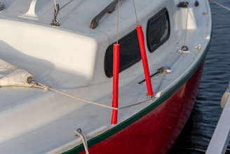 A close-up view of a sailboat with a white deck and red hull, featuring red line protectors hanging on ropes. The boat is situated on calm water, and there are small metal fixtures on the deck.