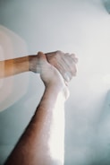 Two hands clasp together in a gesture of support or assistance, illuminated by bright sunlight from above, casting a soft, ethereal glow. The image suggests themes of connection and help.