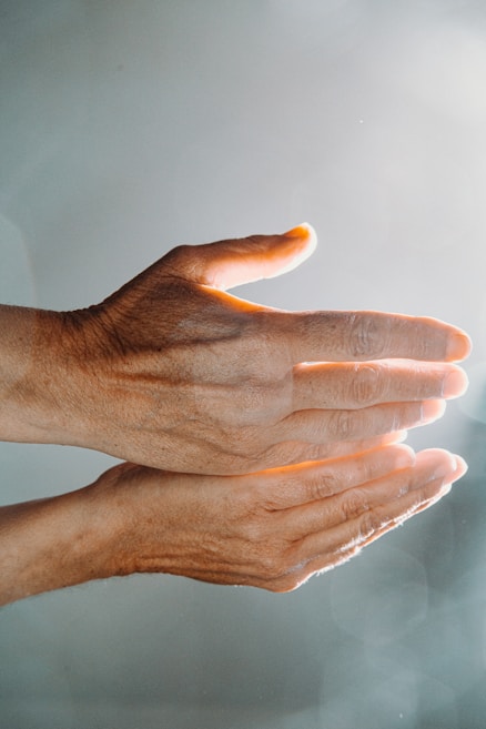 Two hands are positioned in a parallel alignment with fingers slightly spread, bathed in warm, soft light. The light creates a gentle, serene atmosphere and highlights the texture and contours of the skin.