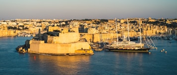 The image captures a historic coastal city bathed in warm sunlight, highlighting the golden tones of the stone architecture. A large yacht is docked in the harbor, surrounded by smaller boats, with the city densely packed along the waterfront. The overall atmosphere suggests a blend of history and modern luxury.