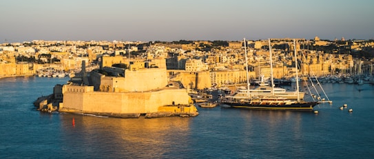 The image captures a historic coastal city bathed in warm sunlight, highlighting the golden tones of the stone architecture. A large yacht is docked in the harbor, surrounded by smaller boats, with the city densely packed along the waterfront. The overall atmosphere suggests a blend of history and modern luxury.