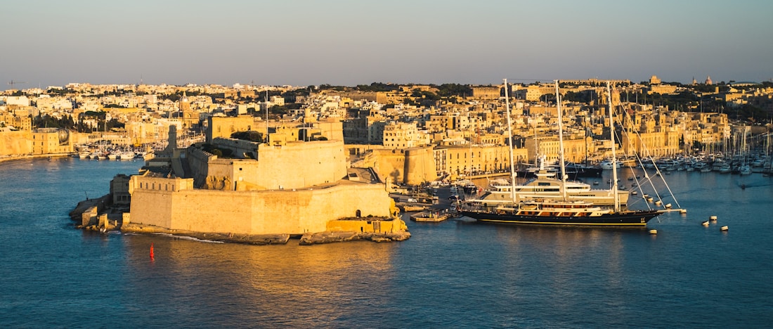 The image captures a historic coastal city bathed in warm sunlight, highlighting the golden tones of the stone architecture. A large yacht is docked in the harbor, surrounded by smaller boats, with the city densely packed along the waterfront. The overall atmosphere suggests a blend of history and modern luxury.