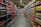 A supermarket aisle with shelves stocked with various beverages, primarily soft drinks and fruit juices. The left side is lined with red and black bottles, while the right side displays cartons of colorful juices. The perspective shows a narrowing view towards the back of the store, lit by overhead fluorescent lights.