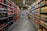 A supermarket aisle with shelves stocked with various beverages, primarily soft drinks and fruit juices. The left side is lined with red and black bottles, while the right side displays cartons of colorful juices. The perspective shows a narrowing view towards the back of the store, lit by overhead fluorescent lights.