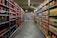 A supermarket aisle with shelves stocked with various beverages, primarily soft drinks and fruit juices. The left side is lined with red and black bottles, while the right side displays cartons of colorful juices. The perspective shows a narrowing view towards the back of the store, lit by overhead fluorescent lights.