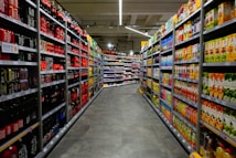 A supermarket aisle with shelves stocked with various beverages, primarily soft drinks and fruit juices. The left side is lined with red and black bottles, while the right side displays cartons of colorful juices. The perspective shows a narrowing view towards the back of the store, lit by overhead fluorescent lights.