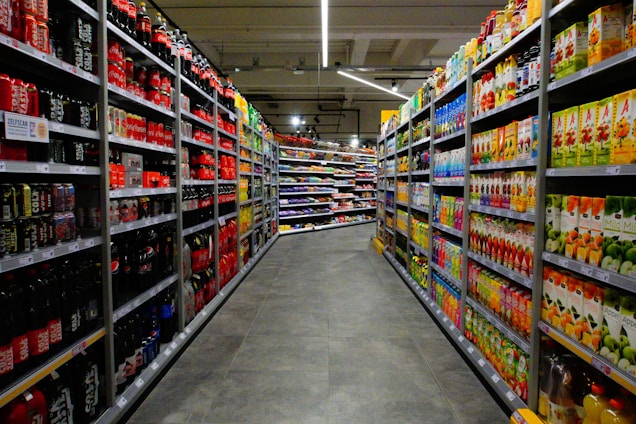 A supermarket aisle with shelves stocked with various beverages, primarily soft drinks and fruit juices. The left side is lined with red and black bottles, while the right side displays cartons of colorful juices. The perspective shows a narrowing view towards the back of the store, lit by overhead fluorescent lights.