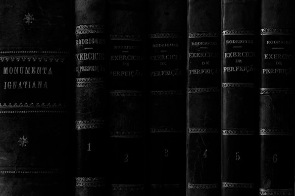 A collection of old, leather-bound books with ornate detailing and faded text is aligned vertically. The titles suggest they are written in Portuguese, with references to exercises of perfection and Ignatian texts. The texture of the leather and the aged appearance of the books convey a sense of history.