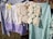 A series of clothes are hanging on a line. Included are a light purple and white skirt with lace detail, a piece of intricate beige crochet, a green checkered cloth, and a blue checkered fabric, all secured with clothespins. The background shows a grassy area with more people and structures.