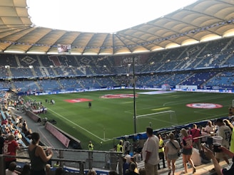 A large football stadium is filled with spectators. The seating area is expansive with blue chairs and features large roof structures providing shade. The well-maintained grass pitch has visible markings and a red circular emblem in the center. People are scattered throughout the stands, with some fans wearing team jerseys, indicating a sporting event atmosphere.