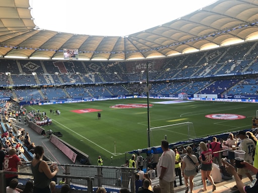 A large football stadium is filled with spectators. The seating area is expansive with blue chairs and features large roof structures providing shade. The well-maintained grass pitch has visible markings and a red circular emblem in the center. People are scattered throughout the stands, with some fans wearing team jerseys, indicating a sporting event atmosphere.
