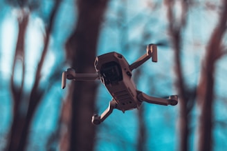 A drone is seen flying in the foreground with blurred trees in the background. The background has a cool, bluish tone while the drone appears sharp and detailed.