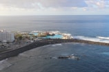 An aerial view of a coastal area with a large hotel building on the left surrounded by palm trees. There is a vibrant blue swimming pool next to the sea, enclosed by a rocky breakwater. The ocean stretches out beyond the pool, with gentle waves crashing against the shore under a partly cloudy sky.