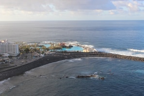 An aerial view of a coastal area with a large hotel building on the left surrounded by palm trees. There is a vibrant blue swimming pool next to the sea, enclosed by a rocky breakwater. The ocean stretches out beyond the pool, with gentle waves crashing against the shore under a partly cloudy sky.