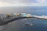 An aerial view of a coastal area with a large hotel building on the left surrounded by palm trees. There is a vibrant blue swimming pool next to the sea, enclosed by a rocky breakwater. The ocean stretches out beyond the pool, with gentle waves crashing against the shore under a partly cloudy sky.