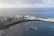 An aerial view of a coastal area with a large hotel building on the left surrounded by palm trees. There is a vibrant blue swimming pool next to the sea, enclosed by a rocky breakwater. The ocean stretches out beyond the pool, with gentle waves crashing against the shore under a partly cloudy sky.