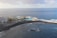 An aerial view of a coastal area with a large hotel building on the left surrounded by palm trees. There is a vibrant blue swimming pool next to the sea, enclosed by a rocky breakwater. The ocean stretches out beyond the pool, with gentle waves crashing against the shore under a partly cloudy sky.