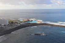 An aerial view of a coastal area with a large hotel building on the left surrounded by palm trees. There is a vibrant blue swimming pool next to the sea, enclosed by a rocky breakwater. The ocean stretches out beyond the pool, with gentle waves crashing against the shore under a partly cloudy sky.