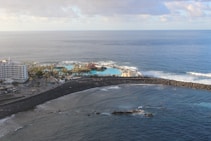 An aerial view of a coastal area with a large hotel building on the left surrounded by palm trees. There is a vibrant blue swimming pool next to the sea, enclosed by a rocky breakwater. The ocean stretches out beyond the pool, with gentle waves crashing against the shore under a partly cloudy sky.