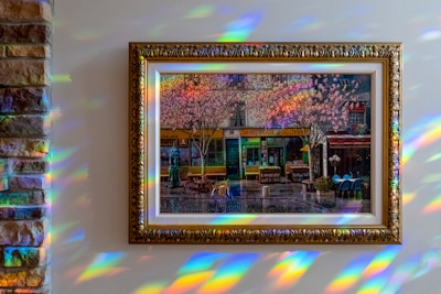 A framed painting of a street scene is displayed on a wall. The artwork depicts a charming urban setting with cherry blossom trees, a bicycle, and quaint shops, including one labeled 'Shakespeare and Company.' The lighting creates a spectrum of rainbow reflections on the wall surrounding the painting.