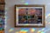 A framed painting of a street scene is displayed on a wall. The artwork depicts a charming urban setting with cherry blossom trees, a bicycle, and quaint shops, including one labeled 'Shakespeare and Company.' The lighting creates a spectrum of rainbow reflections on the wall surrounding the painting.