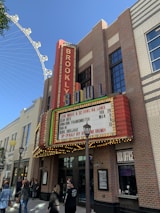 A brick building with a large sign showing 'BROOKLYN BOWL' in colorful neon lights. There is also a marquee advertising live music events and bowling lanes. Various people are walking by or standing in front of the building. Nearby structures include lamp posts and another building with a beige façade. The sky is clear and a Ferris wheel is partially visible in the background.