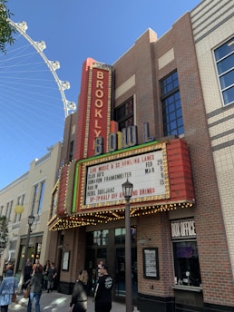 A brick building with a large sign showing 'BROOKLYN BOWL' in colorful neon lights. There is also a marquee advertising live music events and bowling lanes. Various people are walking by or standing in front of the building. Nearby structures include lamp posts and another building with a beige façade. The sky is clear and a Ferris wheel is partially visible in the background.
