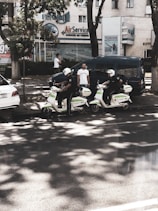 Two individuals wearing helmets and riding scooters are parked on the side of a street. They appear to be part of a law enforcement or security service, as indicated by the markings on their scooters. There are two men standing nearby, one using a phone and another observing the scene. In the background, several buildings feature business signage, and trees provide some shade on the street.